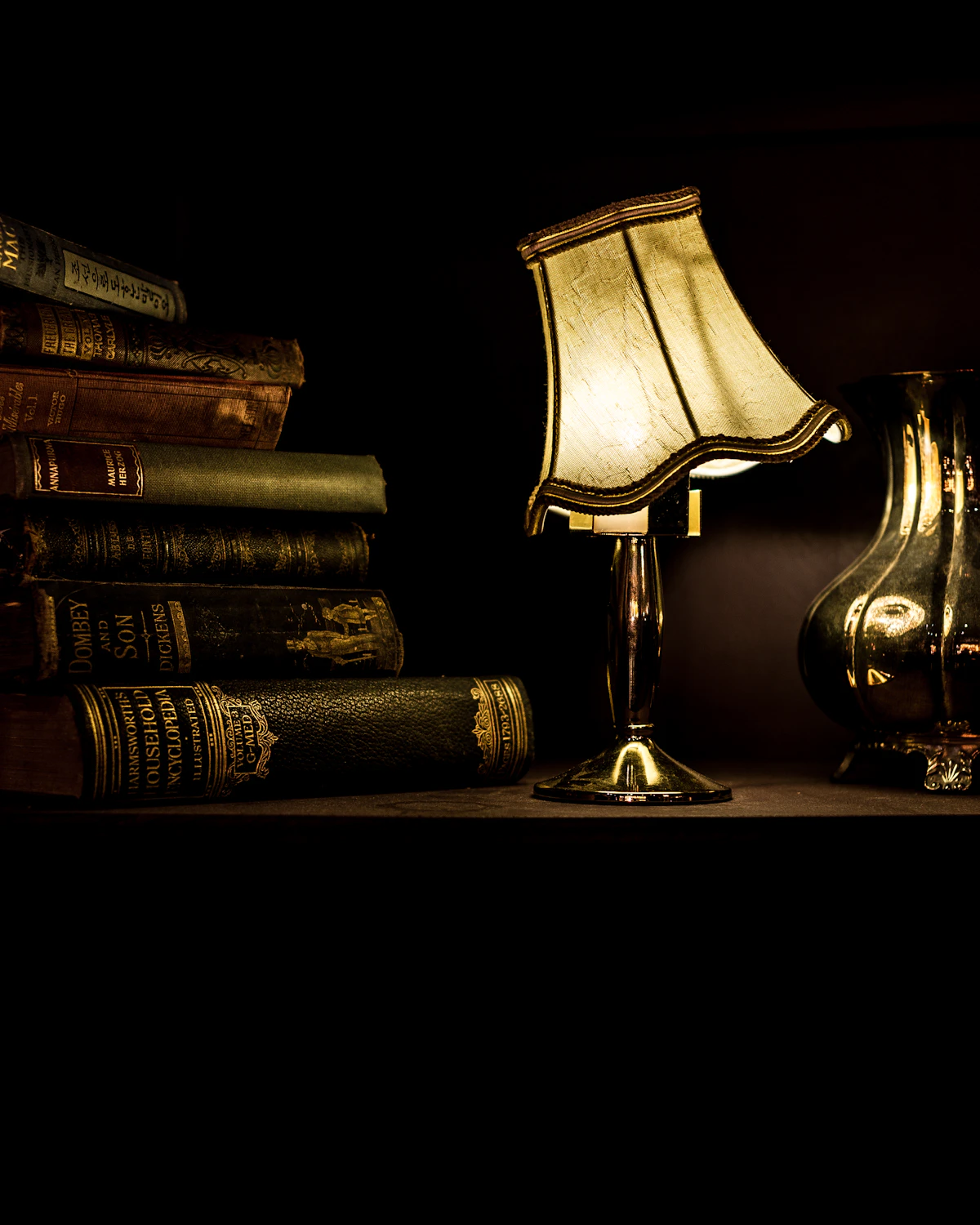 A brass desk lamp lit beside a stack of leather-bound volumes on a dark wood surface.