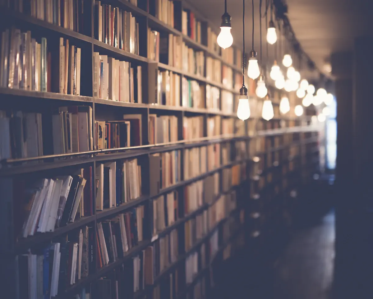 A long library aisle lined with packed bookshelves under a receding row of warm hanging bulb lights.